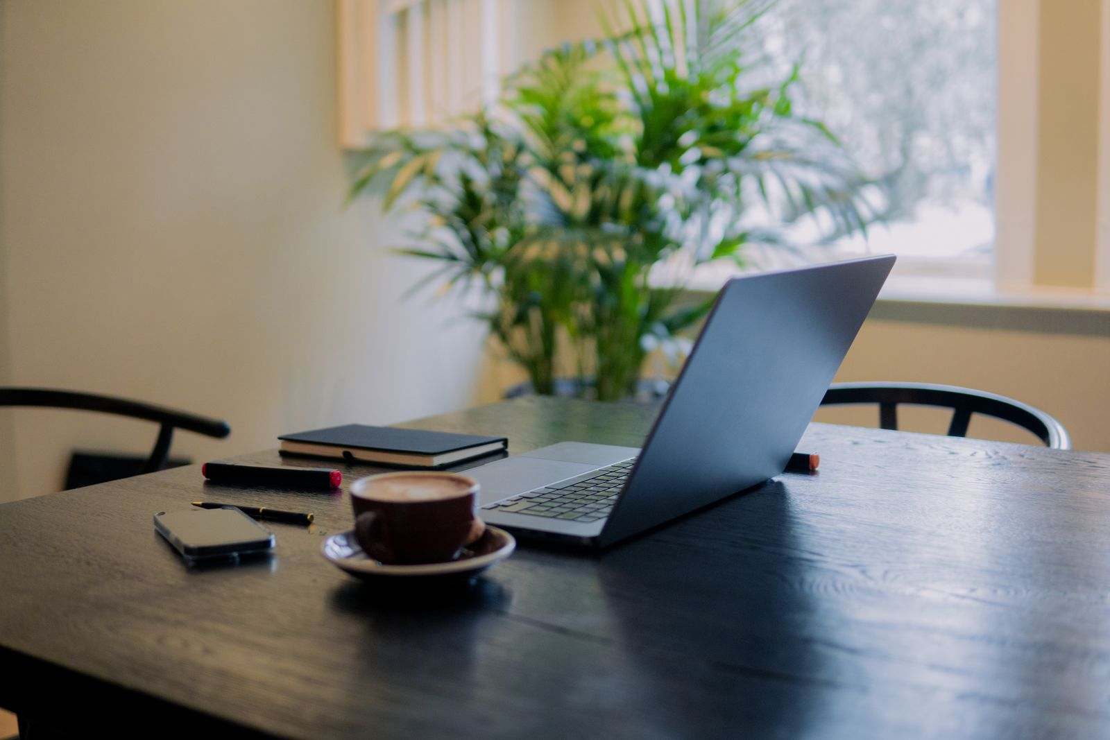 A laptop, coffee, and phone on a dark wooden table by a bright window